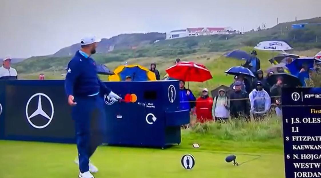 Golfer on a golf course teeing off near spectators with umbrellas.