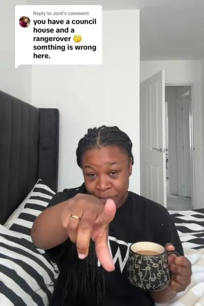 Woman in bedroom holding coffee mug, responding to comment about living in council house and driving Range Rover.