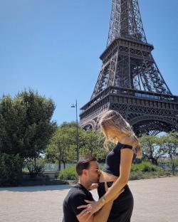 Man kissing pregnant woman's belly in front of the Eiffel Tower.