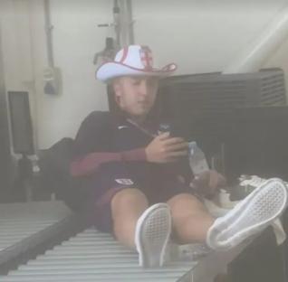 Man in England hat sitting on conveyor belt with drink.