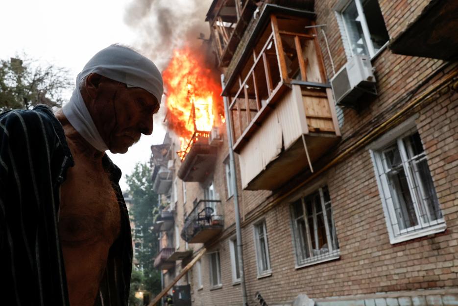 Injured man stands outside burning building.