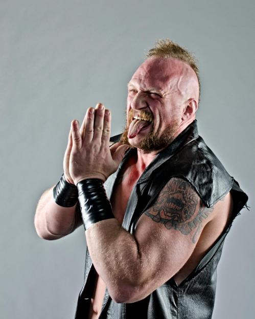A muscular man with a mohawk and tattoos sticking his tongue out.