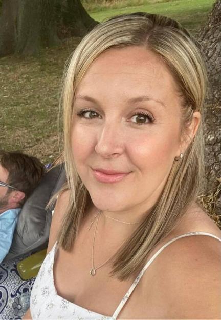 Woman smiling at the camera with a man relaxing behind her, reading a book.