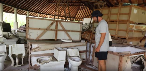 A man stands amidst abandoned, damaged toilets and crates labeled