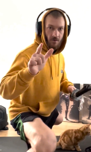 Man in yellow hoodie wearing headphones makes a hand gesture while sitting on the floor with a cat.