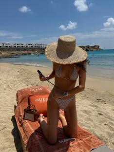 Woman in a bikini kneeling on a beach towel, looking at her phone.