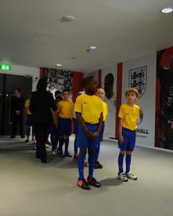 Young soccer players in yellow shirts and blue shorts stand in a hallway with a mural depicting the England national team.