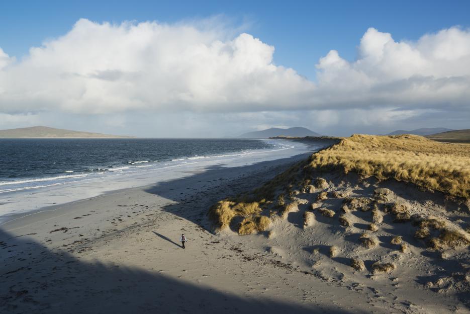 Woman walking on a sandy beach with dunes and ocean in the background.
