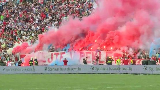 Fans in a stadium holding a banner and setting off red and blue smoke flares.
