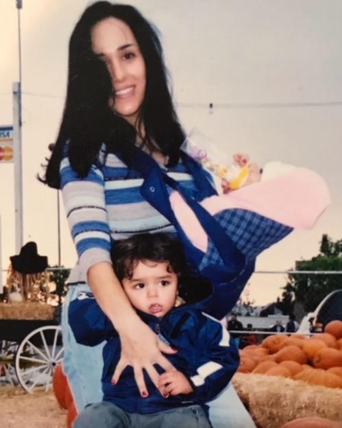 A woman holding two young children in a baby carrier and arms, near pumpkins.