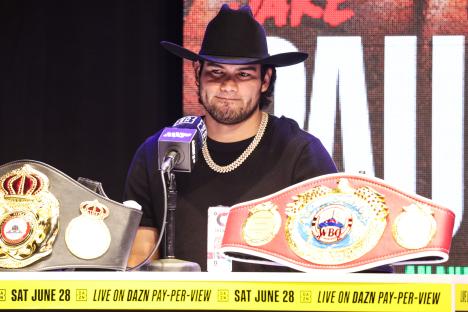 Julio Cesar Chavez Jr. at a press conference, wearing a cowboy hat and gold chain, with boxing championship belts in front of him.