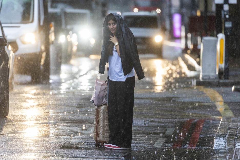 Woman using jacket to shield herself from rain while pulling luggage.