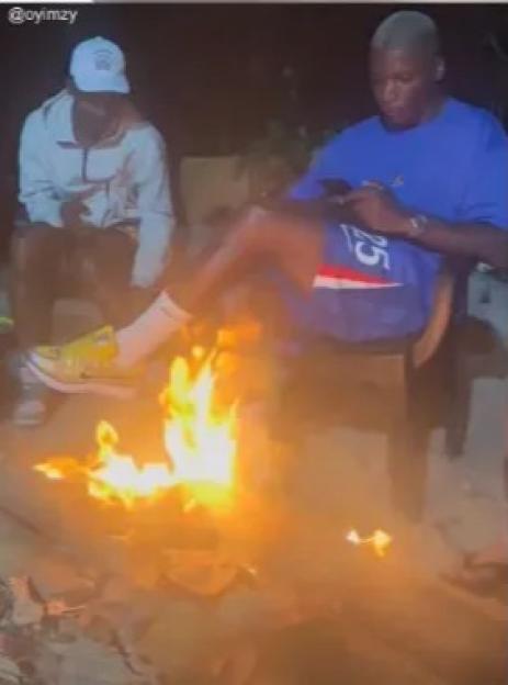 Two men sitting by a bonfire at night, one looking at his phone.