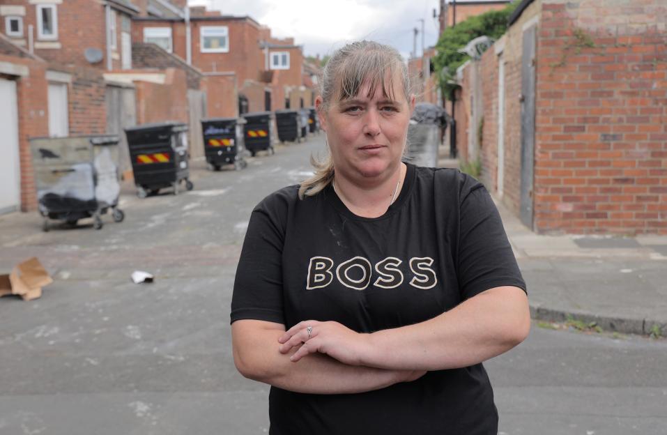 Woman standing in alleyway with rubbish bins in background.