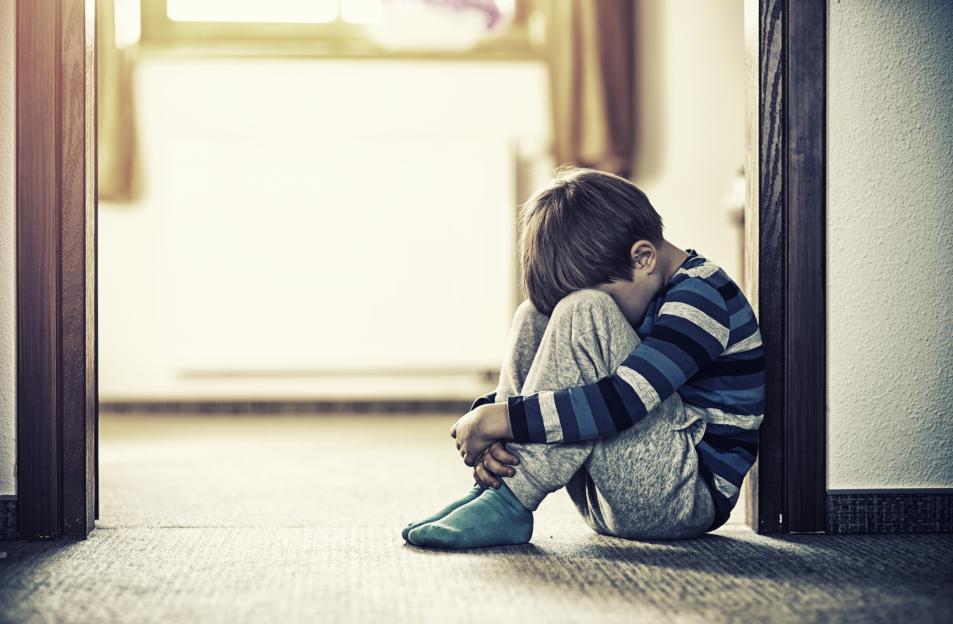 A sad young boy sits on the floor, hugging his knees.