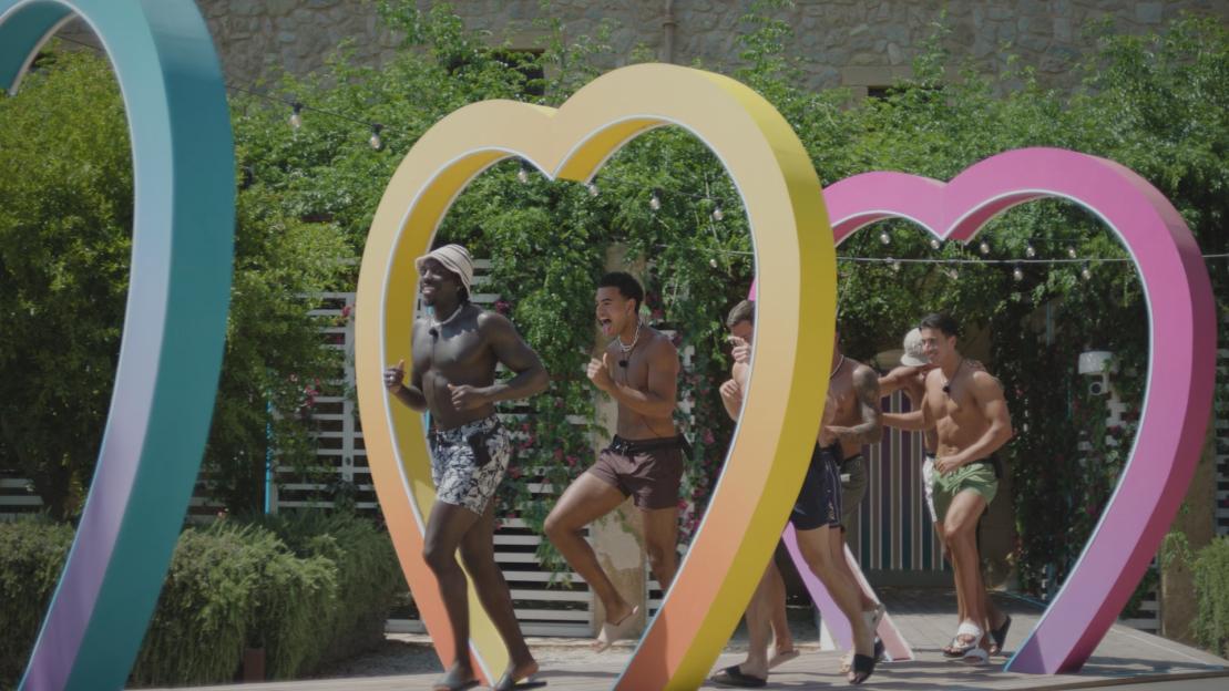 Five men in swim trunks running through heart-shaped arches.