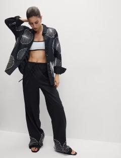 Model wearing a black linen shirt and pants with white shell embroidery.