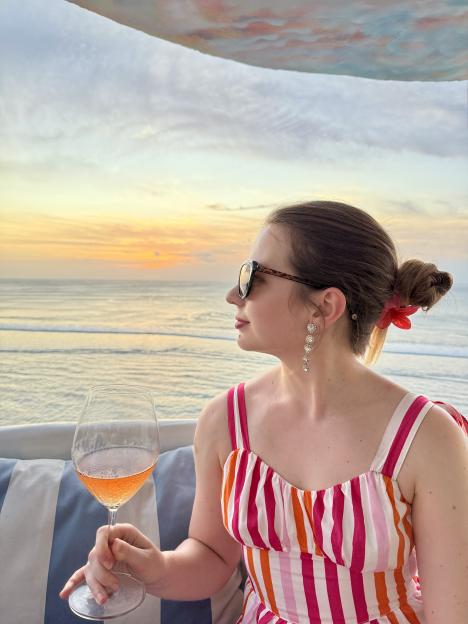 Woman in striped dress enjoying a glass of rosé wine while watching a sunset over the ocean.