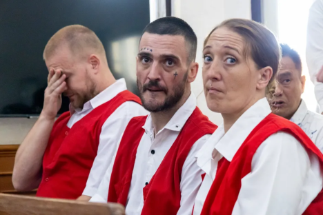 Three people in red vests and white shirts sit, one with their hand on their face.