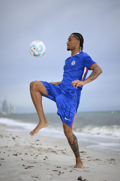 Joao Pedro juggling a soccer ball on a beach in his Chelsea uniform.