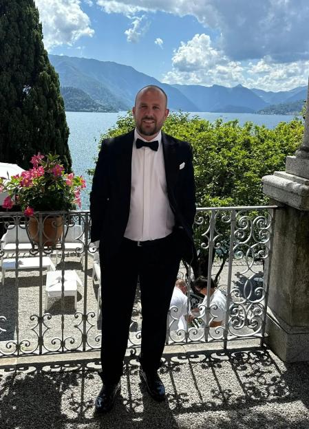 Man in tuxedo on balcony overlooking lake and mountains.