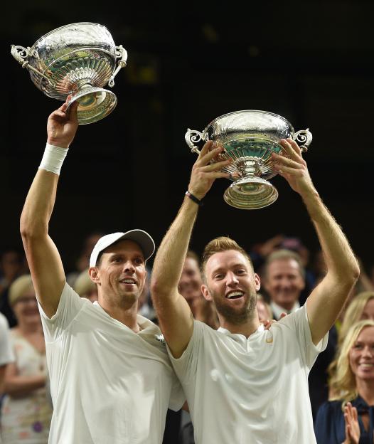 Two male tennis players holding up trophies after winning a match.