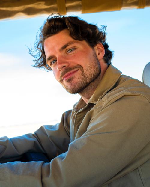 Portrait of a man with wavy brown hair and a light tan jacket.