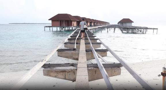 Person walking on a dilapidated pier toward abandoned overwater bungalows.