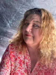 Headshot of a woman with blonde curly hair wearing a pink and white patterned blouse.