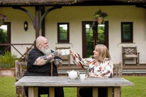A man and woman, sitting at an outdoor table, raise mugs in a toast.