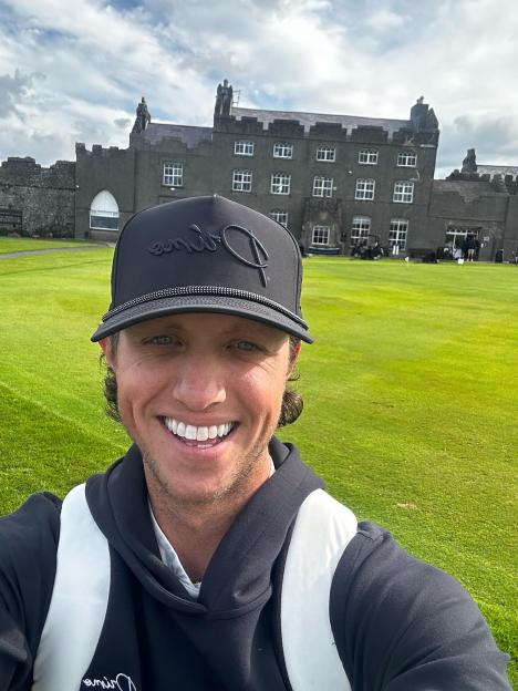 Man in a golf course wearing a black hoodie and cap, with a castle in the background.