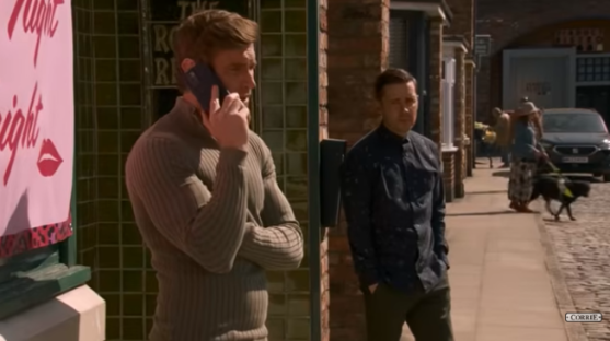 Screenshot of a Coronation Street scene showing two men on a street, one on a phone call.