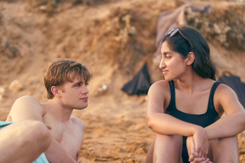 A young man and woman sitting on a beach, looking at each other.
