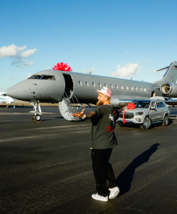 Man standing on tarmac next to a private jet and luxury SUV, both with pink bows.