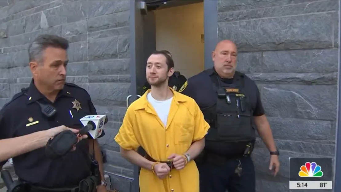 A man in a yellow jumpsuit flanked by two officers is interviewed by a reporter.