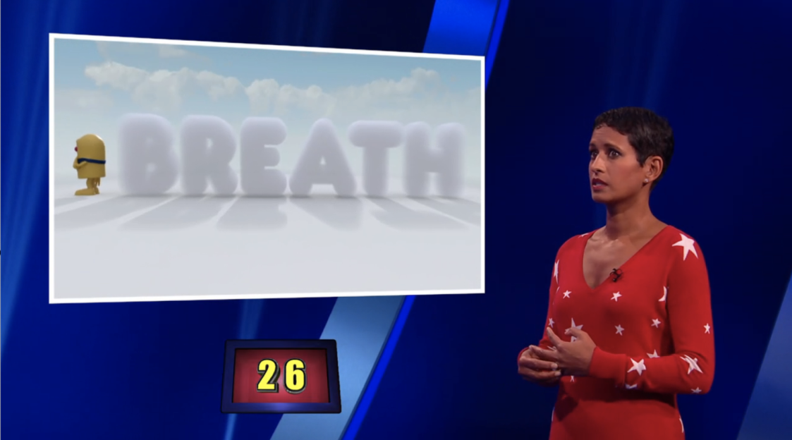 Screenshot of a game show: a woman in a red sweater stands beside a screen displaying the word 