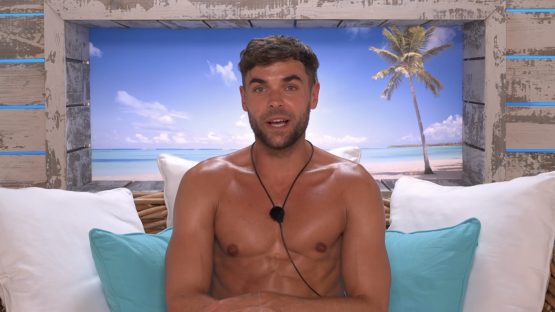 Shirtless man sitting on a couch in front of a beach scene.