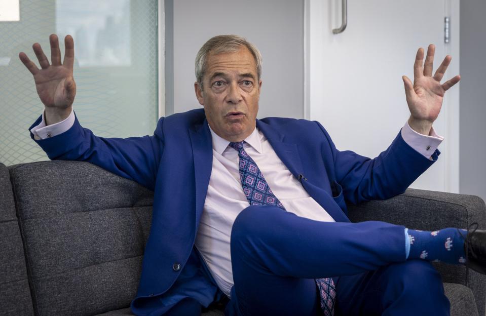 Nigel Farage in a blue suit, sitting on a couch, gesturing with his hands.
