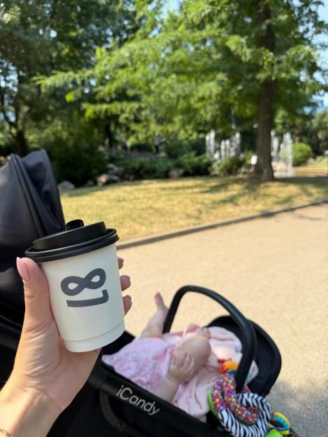 Person holding a coffee cup in a park with a baby in a stroller.