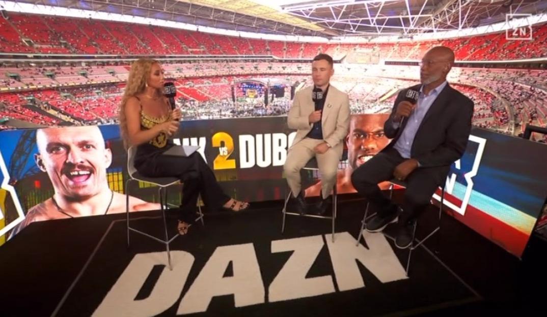 Three boxing commentators sitting in front of a large stadium screen.