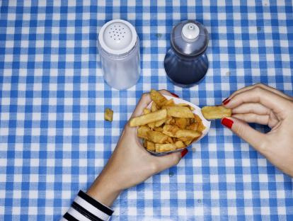Woman's hands holding and eating french fries on a blue and white checkered tablecloth. Salt and vinegar are nearby.