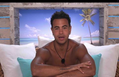 A man sits on a bed, looking at the camera. A tropical beach scene is visible in the background.