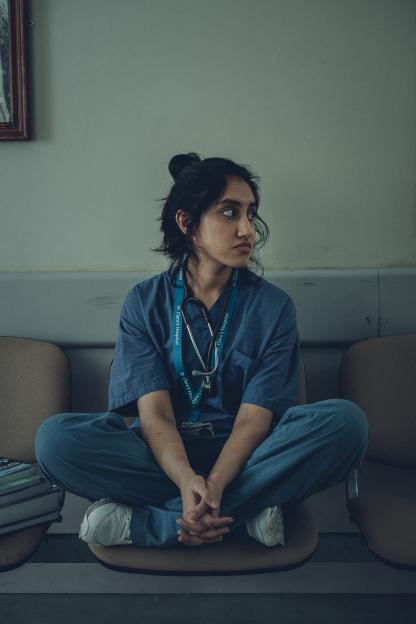 A young woman in scrubs sits cross-legged, wearing a stethoscope.