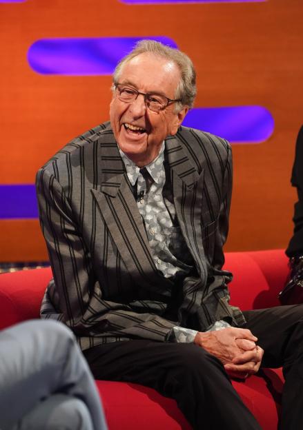 Eric Idle on a red couch, smiling.