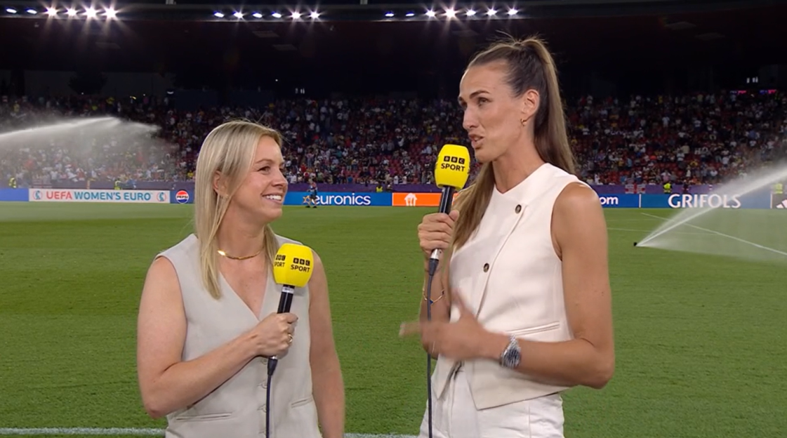 Two women holding BBC Sport microphones conduct an interview on a soccer field.