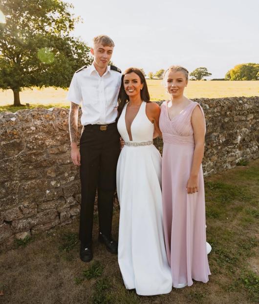 A woman in a white wedding dress stands between a young man in a military uniform and a young woman in a pink dress.