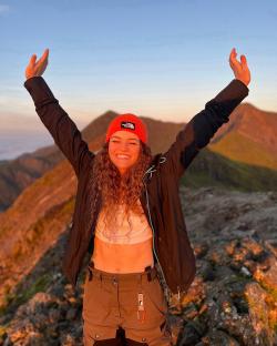 A young woman with arms raised stands atop a mountain at sunset.