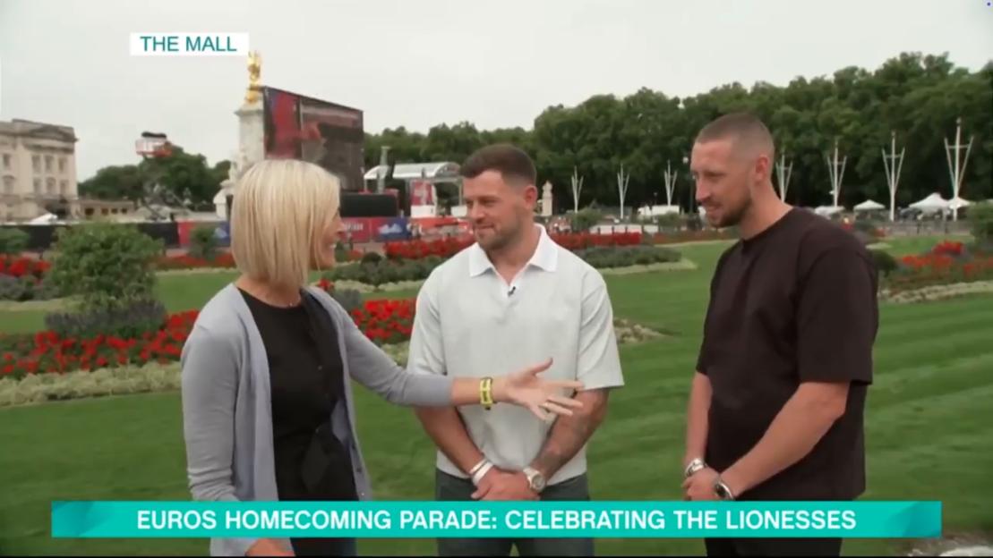 A woman interviewing two men in front of Buckingham Palace during the Euros homecoming parade.