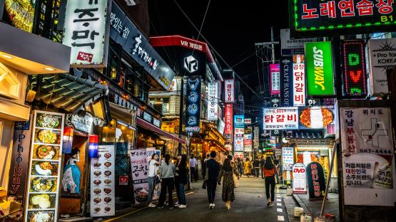 Night view of a bustling pedestrian street in Gangnam, Seoul, with restaurants, shops, and brightly lit signs.