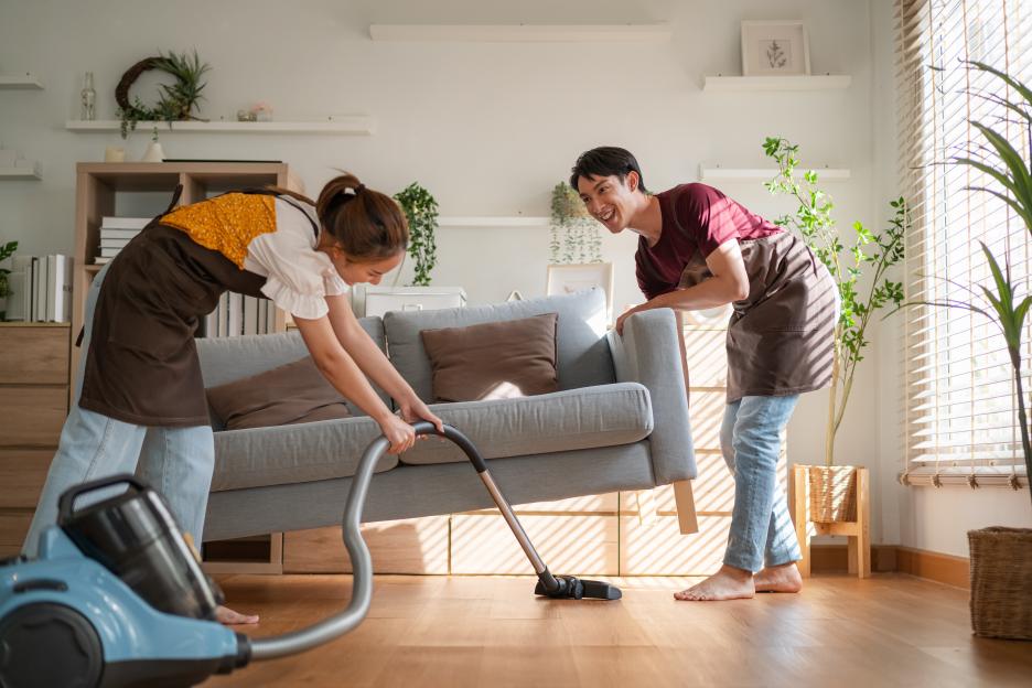 Young couple vacuuming under a sofa in their new home.